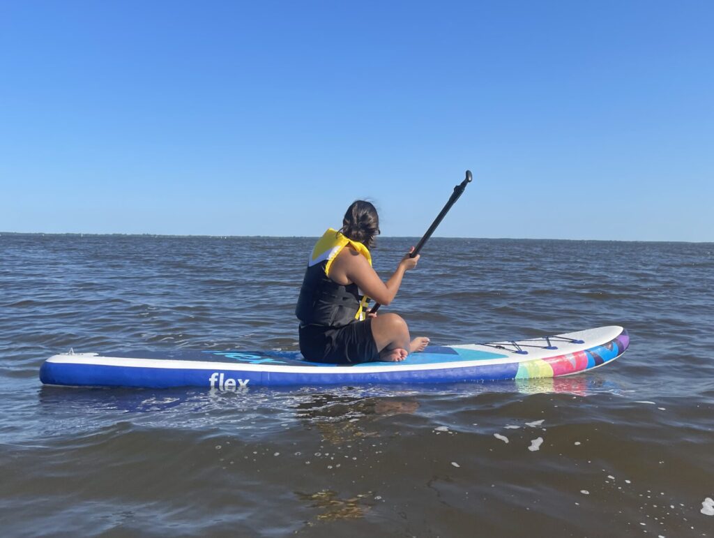 a girl is on a blue paddle board. shes seating down and paddeling the waters. the sky couldn't be any bluer