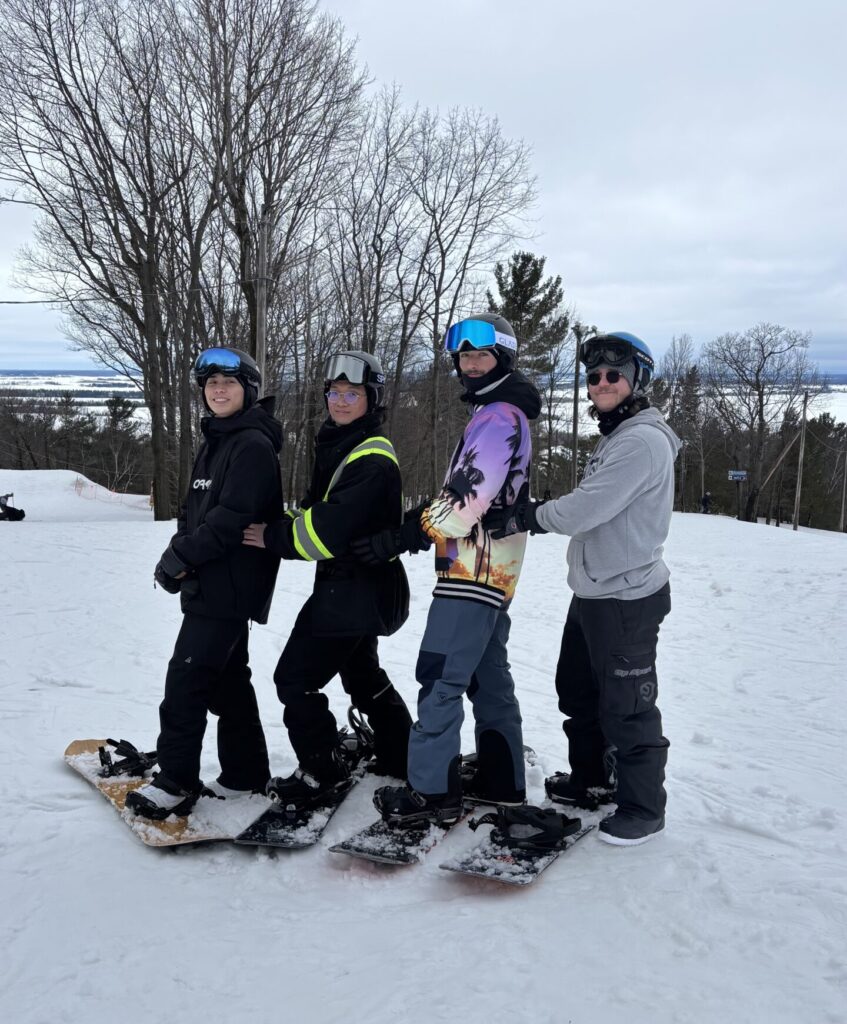 4 guys are posing for a picture. they are all in snowboard gear with one foot on the board. they are all smiling and its a beautiful winter day