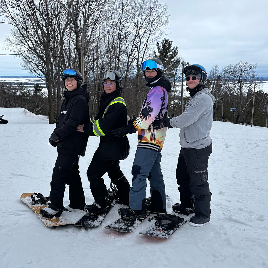 4 guys are posing for a picture. they are all in snowboard gear with one foot on the board. they are all smiling and its a beautiful winter day