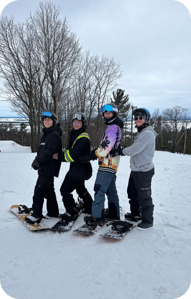 4 guys are posing for a picture. they are all in snowboard gear with one foot on the board. they are all smiling and its a beautiful winter day